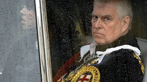 Reuters Prince Andrew sitting behind a glass window in a car with raindrops on it. He is looking glum and wearing a royal uniform with golden trim and a St George's cross on it.