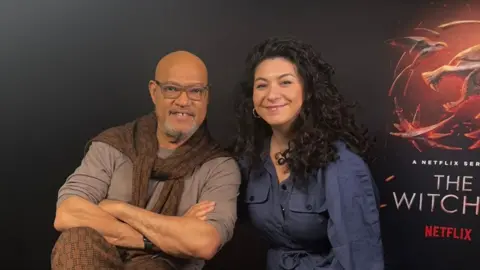 Sabrina Barr Laurence Fishburne crosses his arms and is wearing a grey top and has a brown sweater knotted around his neck. He has a grey goatie and is wearing glasses. Sabrina is to his left with long curly black hair. She's wearing a blue jump suit. Behind them is a promo poster for The Witcher.
