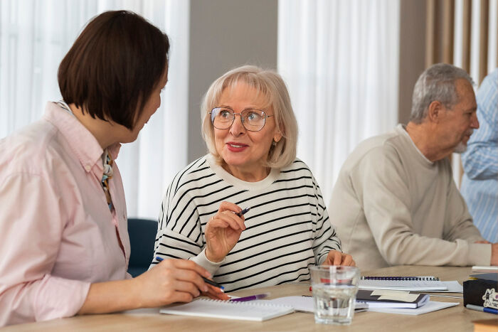 Elderly woman with dementia sharing unfiltered and funny thoughts during a group conversation at a table.