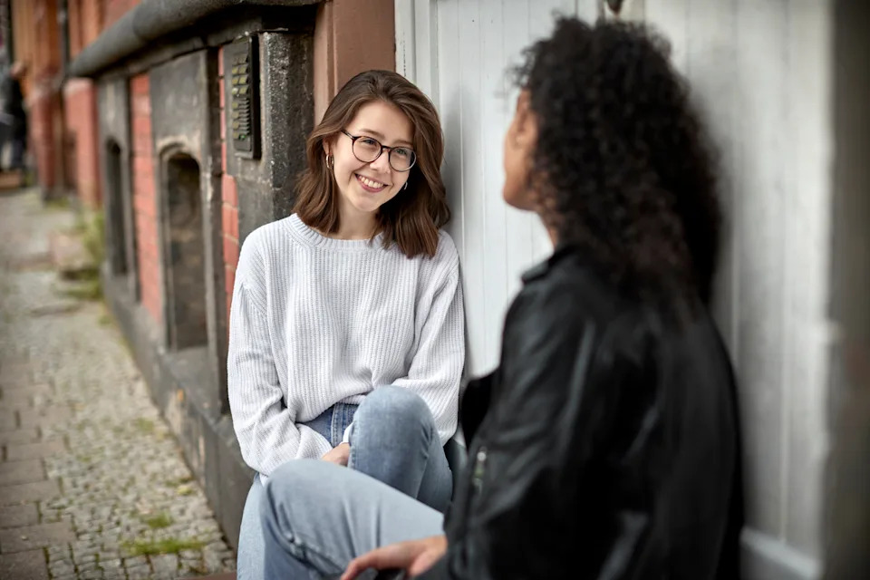 Two people sit on a sidewalk talking, one wears glasses and a casual sweater, the other has curly hair and a leather jacket, both appear engaged