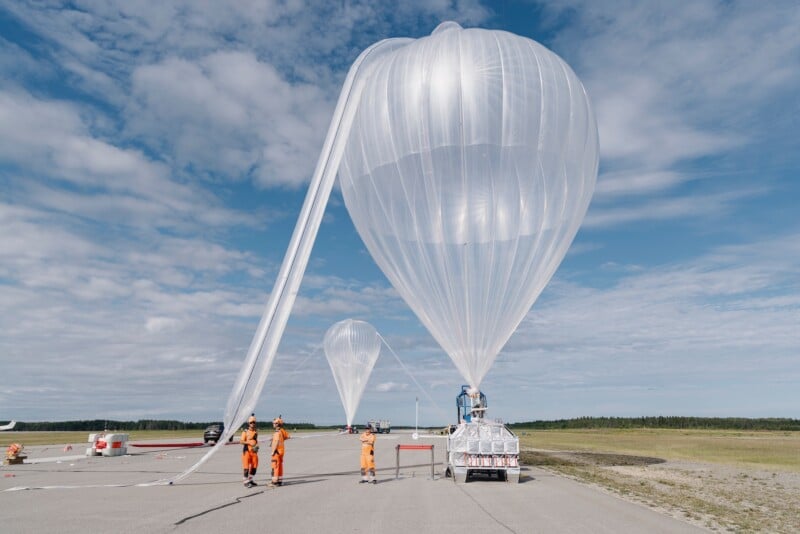 Several people in orange suits prepare large, transparent weather balloons for launch on a wide runway under a partly cloudy sky. One person works on a platform next to a balloon, and more balloons are visible in the background.