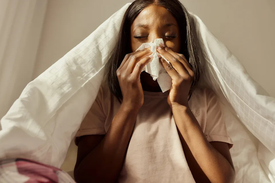 Woman sitting under a blanket, visibly sick, blowing her nose with a tissue