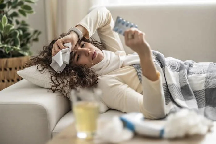 Person lying on a couch looking unwell, holding medication, with tissues and a drink nearby. A checkered blanket is partially covering them