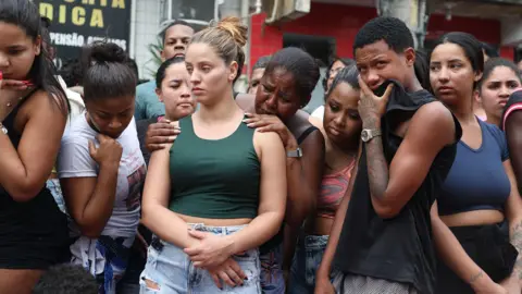 Bruno Itan A group of people - many of them women - look at the ground where bodies have been placed. One man is covering his mouth with his T-shirt. A woman is grabbing the shoulders of the woman in front of her and is crying.