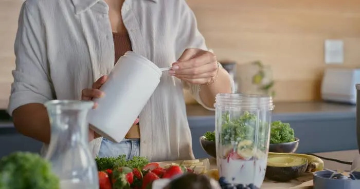 Person adding a scoop of powdered supplement into a blender filled with fruits and greens, preparing a healthy smoothie in a kitchen setting