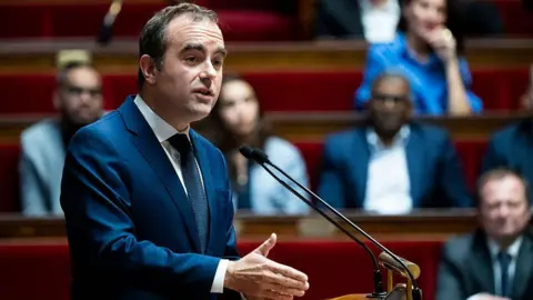 AFP via Getty Images A man in a suit talks in parliament in front of a microphone with MPs looking on in the background
