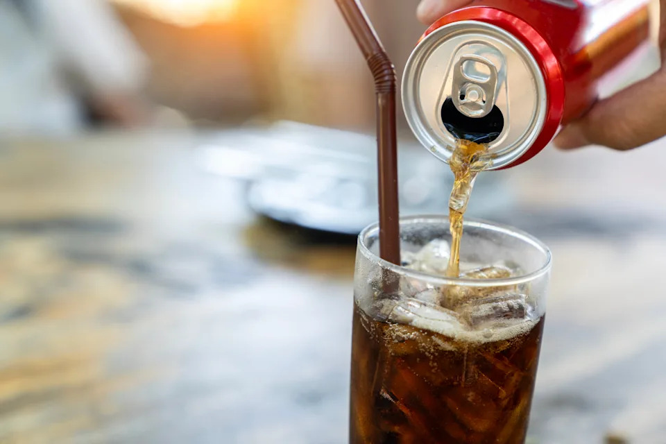 Soda being poured from a red can into a glass with ice and a brown straw, on a blurred table setting