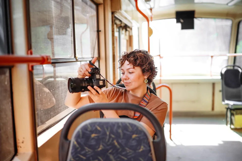 A person on a bus is adjusting a camera, focusing on an outside view through the window