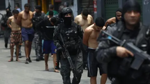 EPA/Shutterstock Rio de Janeiro Police officers guard a group of people during an operation in Rio de Janeiro. The officers are carrying weapons and one of them is wearing a balaclava. The men they are escorting have been handcuffed.
