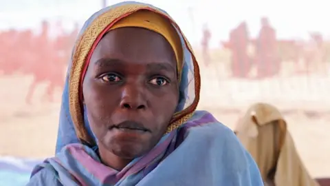 A head and shoulders shot of a woman in a blue headscarf. The gauze of a tent can be seen behind her and some figures are out of focus.