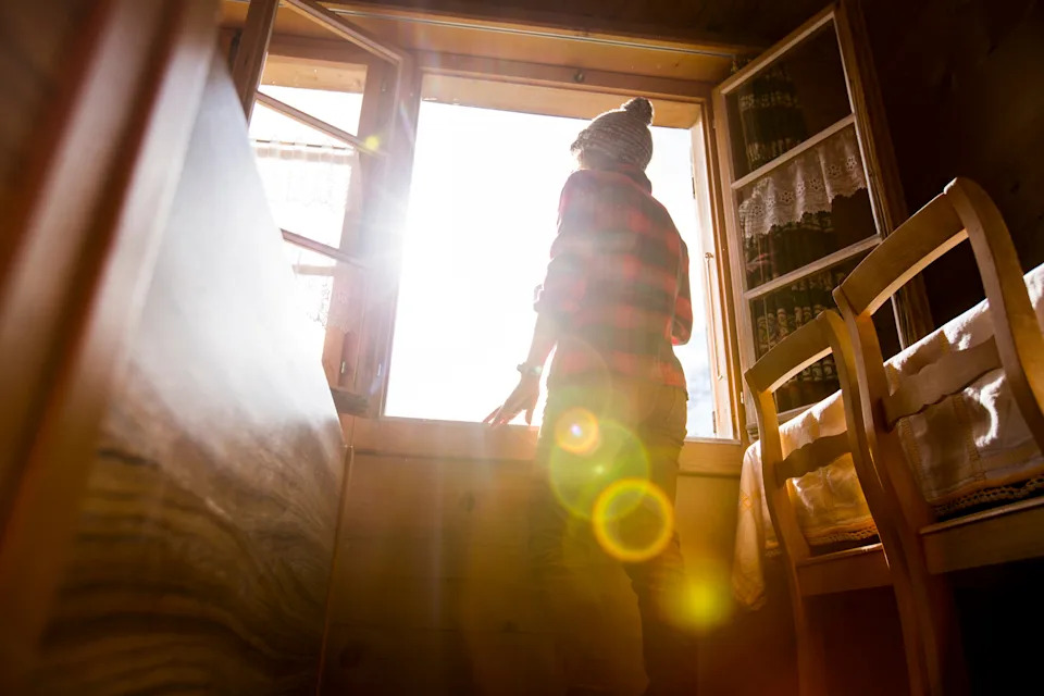 Person in winter clothing stands by an open window, sunlight streaming in, creating a warm indoor atmosphere. A chair and books are nearby