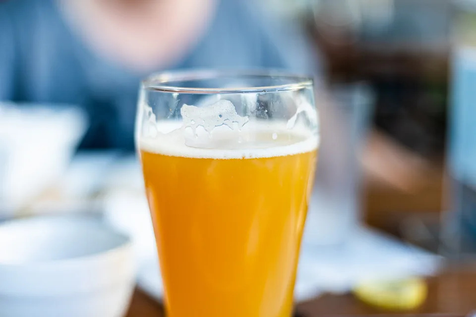 A glass of frothy, light beer on a table in a casual dining setting, with blurred background elements