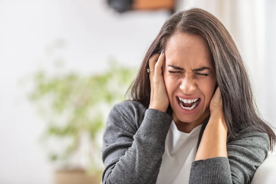 A person holds their head with both hands, eyes closed, appearing stressed or overwhelmed, in a home setting