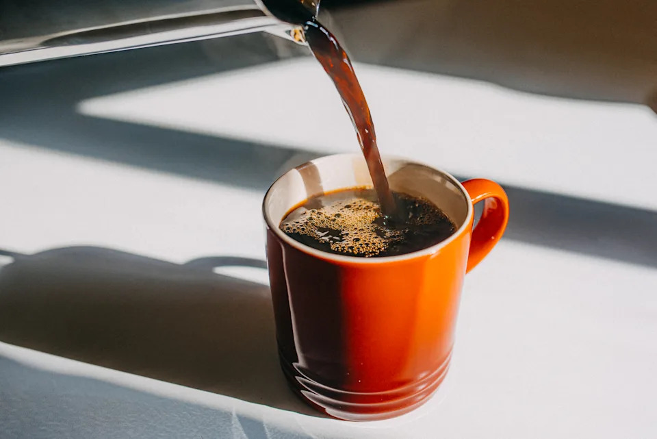 Coffee being poured into a red mug on a table with sunlight filtering through a window