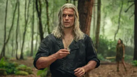 Netflix Henry Cavill as Geralt with long grey hair and cuts all over his face. He holds a silver pendant. He's wearing an open-buttoned green shirt with sleeves rolled up. He's in a forest.