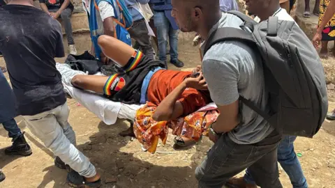BBC An injured man, in a brown shirt and black short, is carried on a stretcher