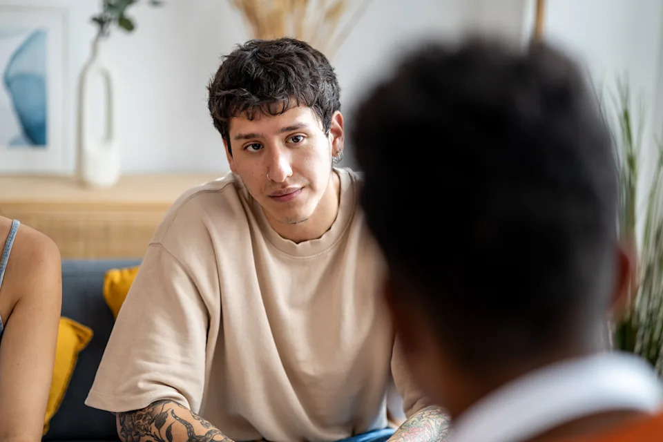 Person with tattoos and short hair attentively listens to someone off-camera in a casual setting with soft focus background decor