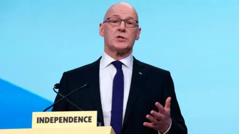 PA Media John Swinney, standing behind a podium labelled Independence and gesturing with his left hand, delivers his keynote speech at the SNP annual conference in Aberdeen