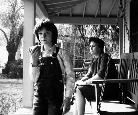 Getty Images Actress Mary Badham, short pudding bowl styledark hair eating an apple & dressed in dungarees and Harper Lee seated on swing bench in porch on the set of the film "To Kill A Mockingbird"