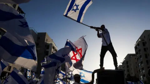 Reuters A man waves an Israeli flag as family and supporters gather on the day that former Israeli hostage Elkana Bohbot returned home after leaving hospital, six days after his release from captivity in Gaza, in Mevaseret Zion, Israel (19 October 2025)