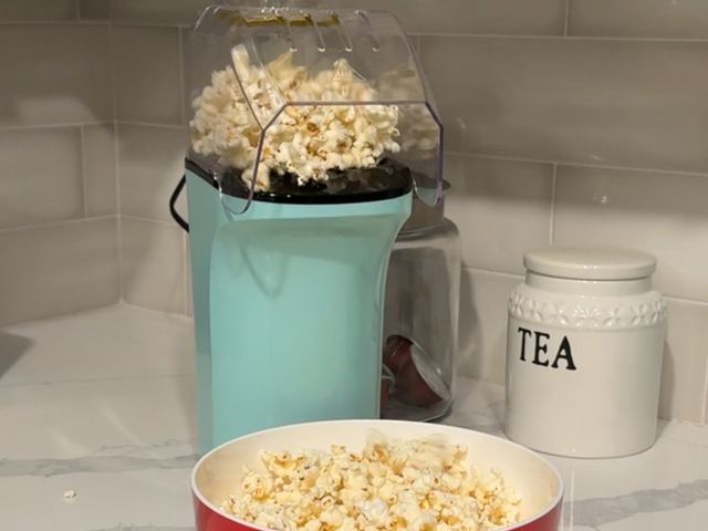 Dash Popcorn Maker, a rectangular appliance with a teal base and clear top with popcorn spilling from the spout into a clear bowl