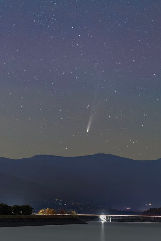 A bright comet with a visible tail streaks across a star-filled night sky above dark, rolling hills. Below, a well-lit bridge crosses a calm river, with some trees and lights along the shore.