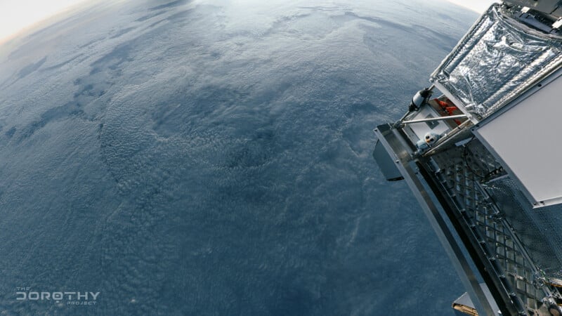 A view of Earth’s curved horizon from near space, showing a textured cloud layer over the blue surface. Part of a metallic scientific instrument or satellite is visible on the right side. “The Dorothy Project” logo is in the corner.