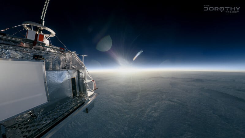 A high-altitude photo shows the edge of a metallic scientific instrument at the left with Earth’s horizon and clouds below, illuminated by the sun in a dark blue sky. The Dorothy Project logo appears in the corner.