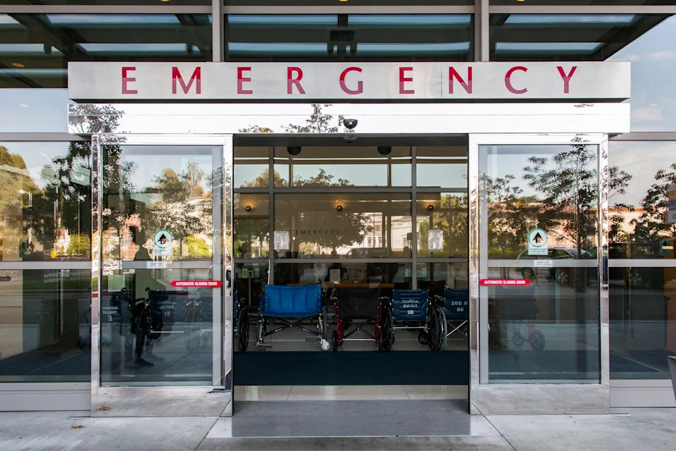 Hospital entrance with glass doors labeled "Emergency," featuring visible wheelchairs inside