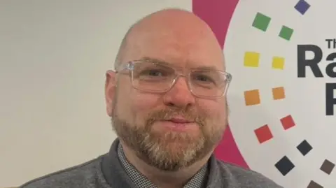 A man with a bald head and beard wearing square glasses looking into the camera. He is wearing a grey jacket and a purple tie. He is standing in front of a sign that says the rainbow project, which is mostly pink but has different colours.