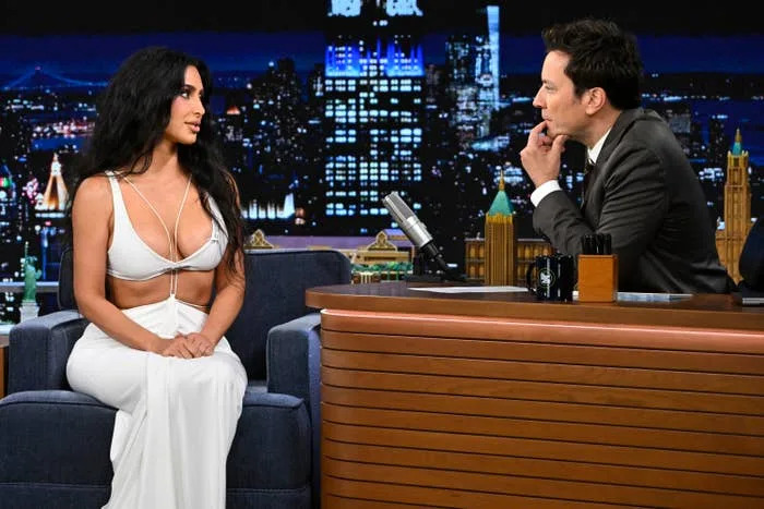 Kim Kardashian in a stylish cut-out dress sits on the Jimmy Fallon talk show set, engaged in conversation