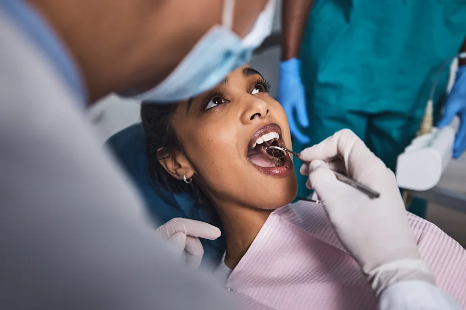 Person at a dental check-up, seated, with a dentist in gloves examining their teeth using a dental mirror and probe