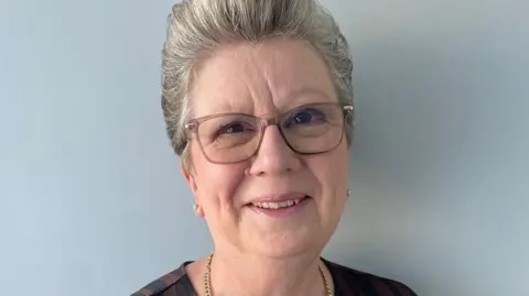 Deena Banister Deena Banister with short grey hair and brown eyes wearing brown glasses and a brown top. She is smiling.