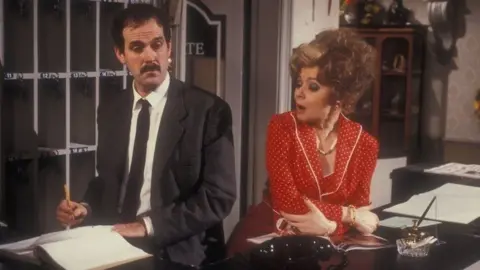 John Cleese & Prunella Scales in Fawlty Towers.  They are shown arguing in the hotel lobby behind the front desk.