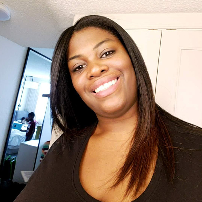 Smiling woman with long straight hair in a black top, with a mirror reflecting a child sitting on a bed behind her.