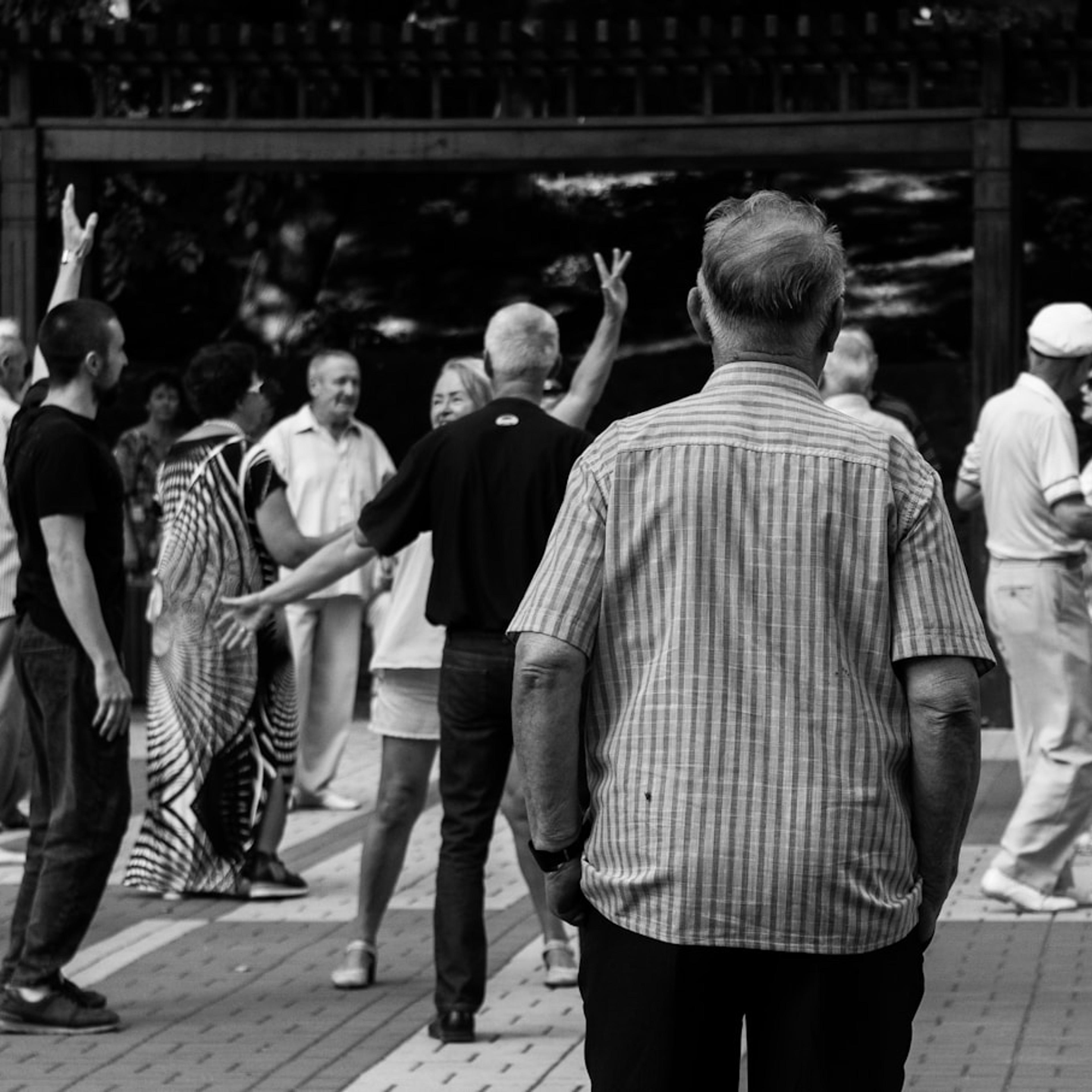Black and white photo of a group of people dancing outdoors, with a man in a striped shirt in the foreground.