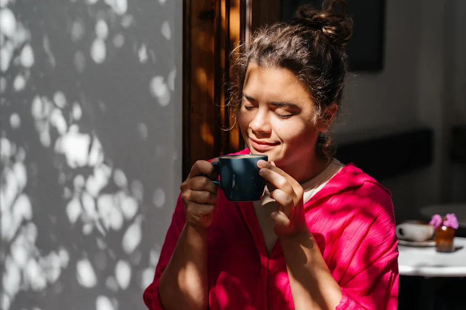 Person in a relaxed outdoor setting, wearing casual clothing, enjoying a cup of coffee with a peaceful smile. Shadows of leaves create a dappled light