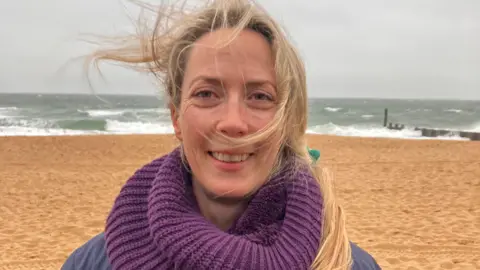 BBC Nicky on Bournemouth beach she has a blue coat and a purple snood on. She has long blond hair which is blowing around in the breeze. The sea is grey and has crashing waves behind her.
