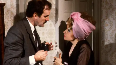 John Cleese and Prunella Scales as Basil and Sybil Fawlty in the 1970s. This is a scene from Fawlty Towers.  Prunella Scales is wearing curlers and a hair cloth and she is arguing with John Cleese.