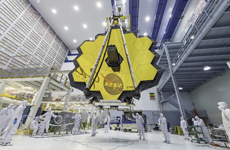 NASA technicians lift the James Webb Space Telescope using a crane and move it inside a clean room.