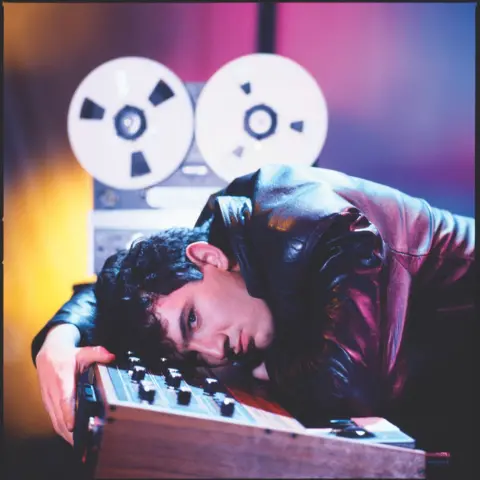 Peter Ashworth Dave Ball is seen lying on top of a 1980s synthesizer, with a reel-to-reel tape machine in the background