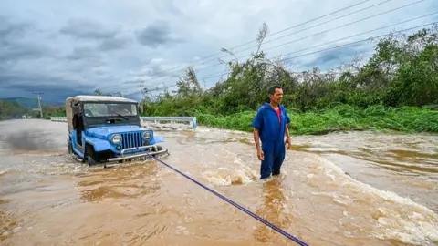 AFP via Getty Images A man wearing a blue jumpsuit stands in brown floodwater beside a bright blue car that is partially submerged. The vehicle is being pulled or secured with a rope attached to the front bumper.
