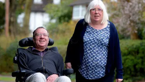 UK MND Research Institute A man (David Setters) in a wheelchair alongside his wife who is his principal carer, in a park. They are both smiling. There are trees and hedges behind them.