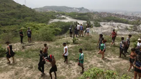 Bruno Itan Around two dozen residents of Penha search a hillside for people who went missing after a police raid. Some of them are looking down what looks like a ravine, while others are walking.