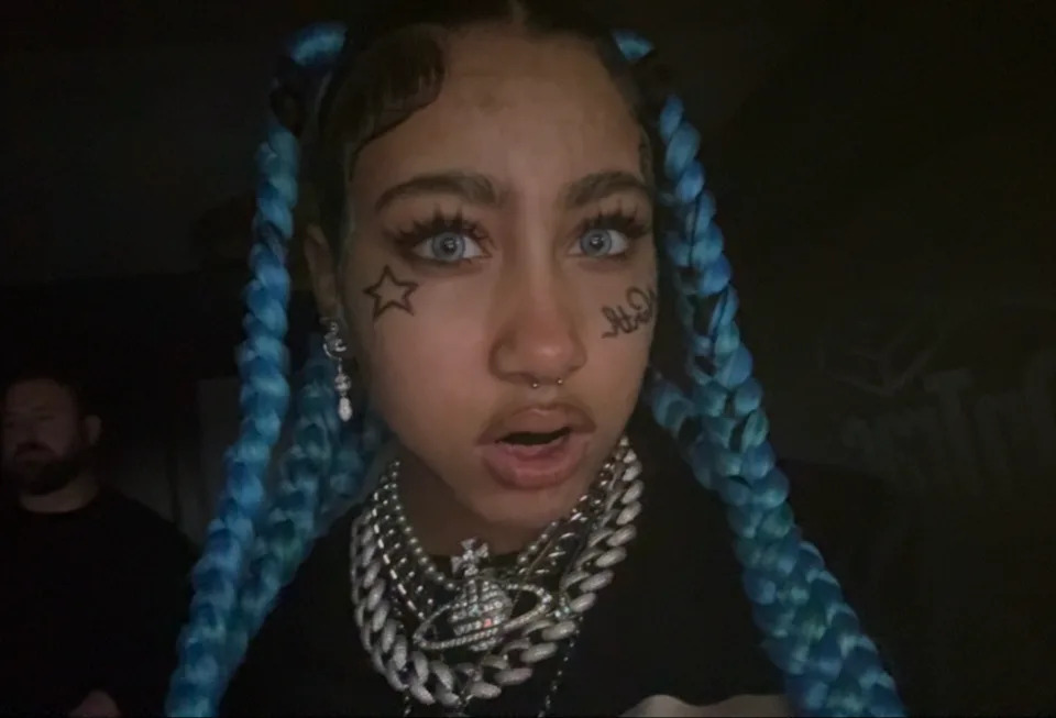 North West with braids and prominent faux face tattoos, wearing layered necklaces, looking directly at the camera
