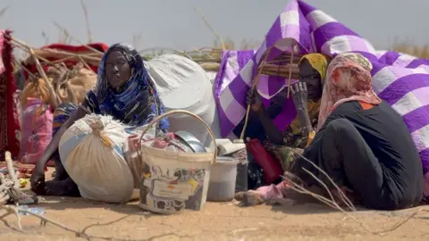Three women wrapped in headscarves sitting on the ground in front of makeshift tents. Some of their belongings in buckets and bags lie in front of them.