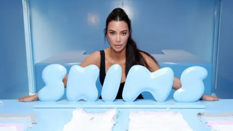 Getty Images Entrepreneur Kim Kardashian poses for a picture while positioned behind a sky-blue Skims logo propped up on the side of a blue table with her company's clothes.