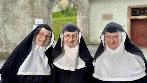 Bethany Bell/BBC Three nuns stand in front of the monastery in their habits, with Sister Rita on the left and Sister Regina in the centre both wearing glasses
