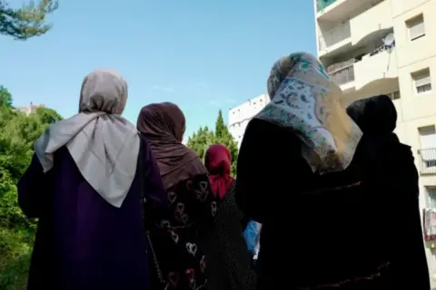 VALERY HACHE/AFP via Getty Images A group of five women in headscarves with their backs to the camera stand in an urban area with greenery on the left side and multi-story residential buildings on the right