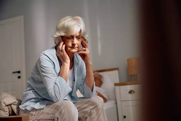 Senior woman holding her head in pain while sitting with eyes closed on the edge of her bed